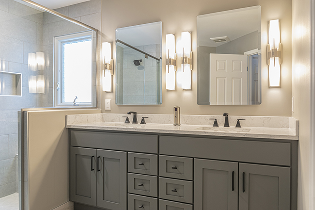 Grey solid wood vanity with a quartz countertop and dual mirrors right next to a walk in tile shower.