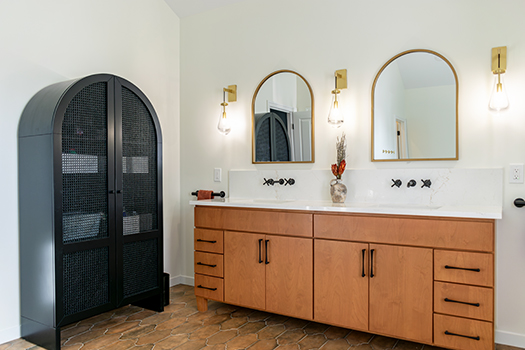 Contemporary bathroom design featuring a wooden double sink vanity, twin arched gold-trimmed mirrors, and elegant wall-mounted gold light fixtures, complemented by a tall black mesh cabinet to the left.