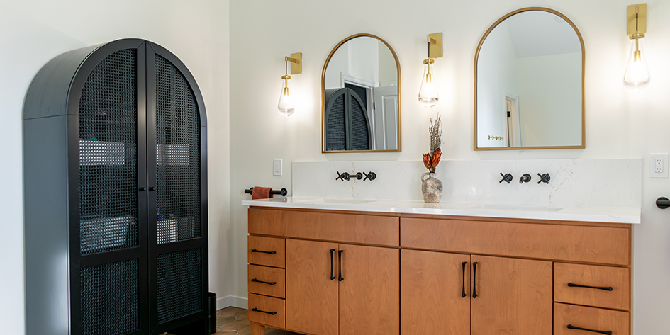 Modern bathroom interior with double vanity cabinet in natural wood, white countertop, two arch mirrors with brass frames, and brass sconces. A tall black armoire with wire mesh doors stands to the left.